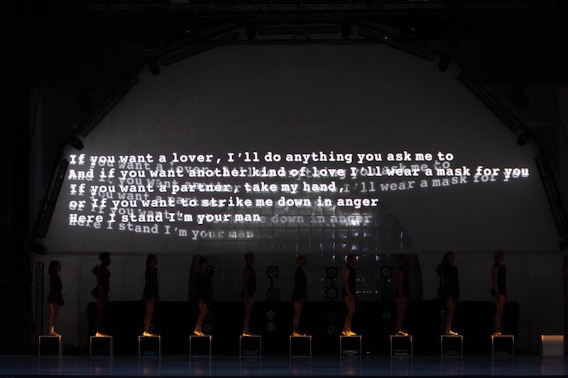 Leonard Cohen lyrics are projected on a back wall while dancers stand atop boxes in a single file line.